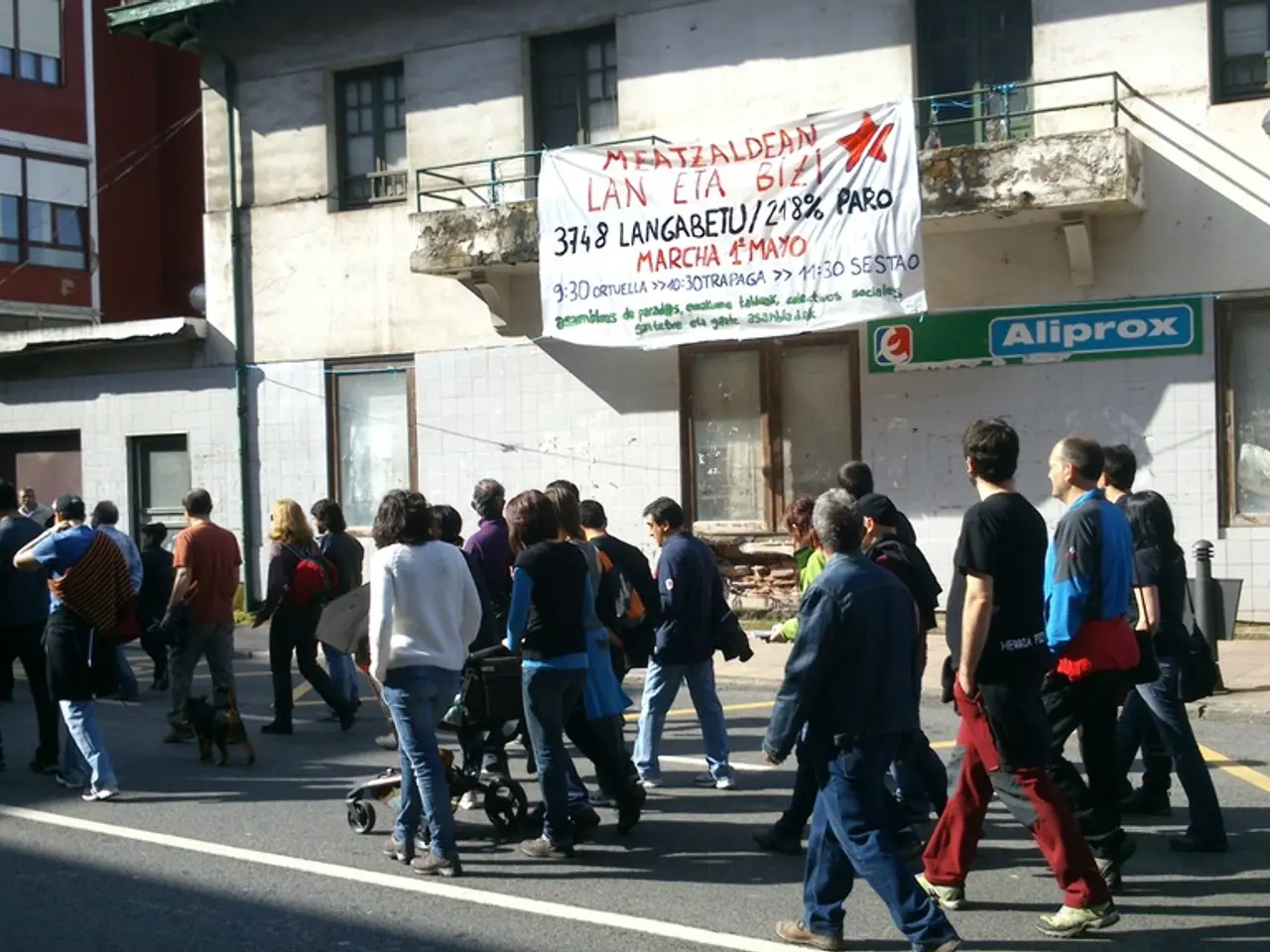 In this image, we can see buildings. There are persons walking on the road. There is a banner at...