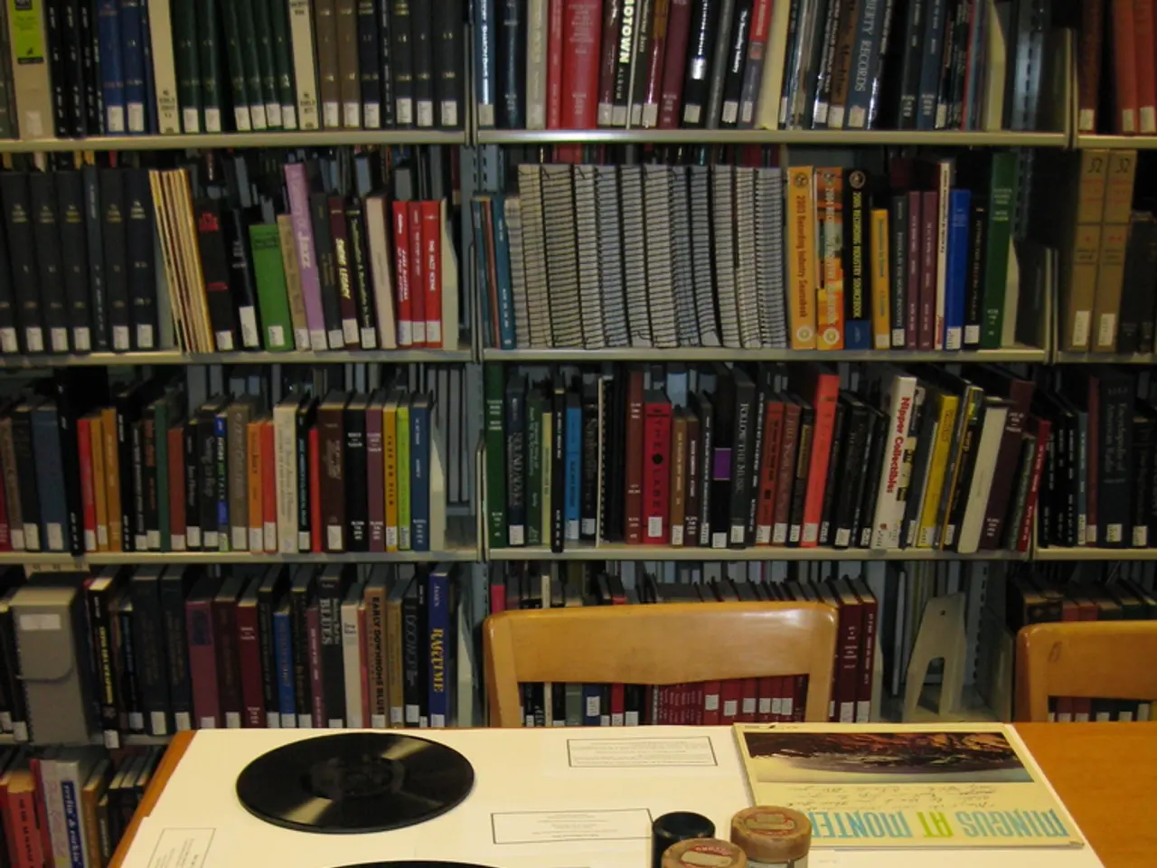 In this image I can see few Cd's and few objects on the table. Back I can see chairs and book-rack....