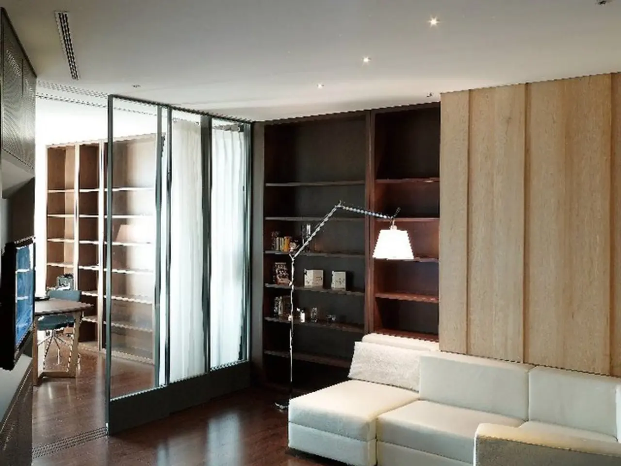 In this picture we can see a room with sofa, light, racks, curtains, table, chair, wooden floor.