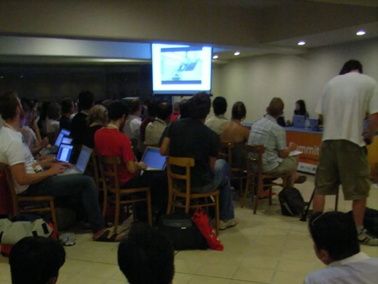 This image is clicked inside the hall. In this image there are few people sitting in the chairs and...