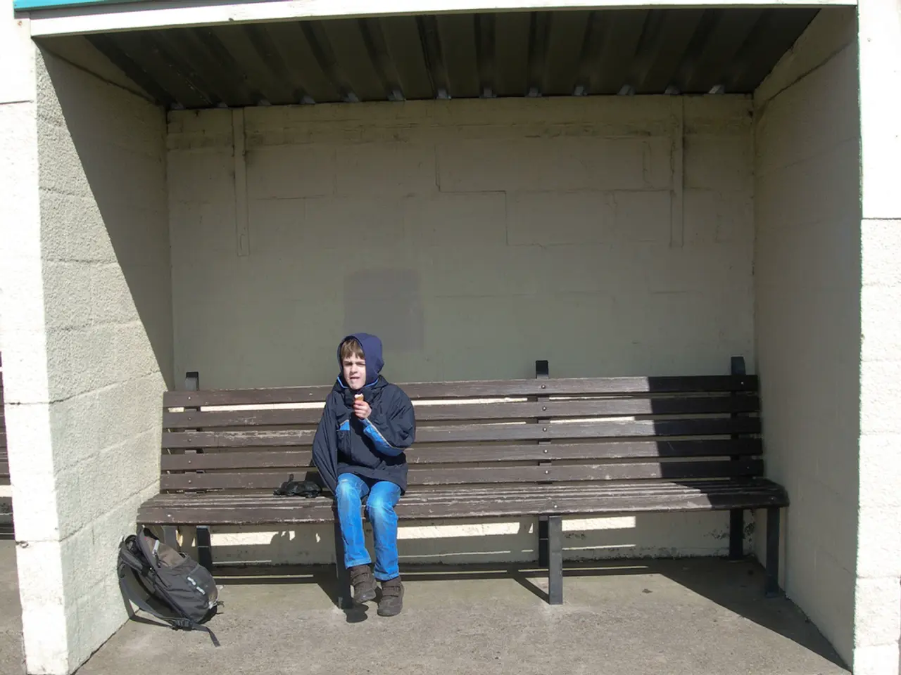 In the image there is a bus stop and there is a bench on which a boy sat with a hoodie and there is...