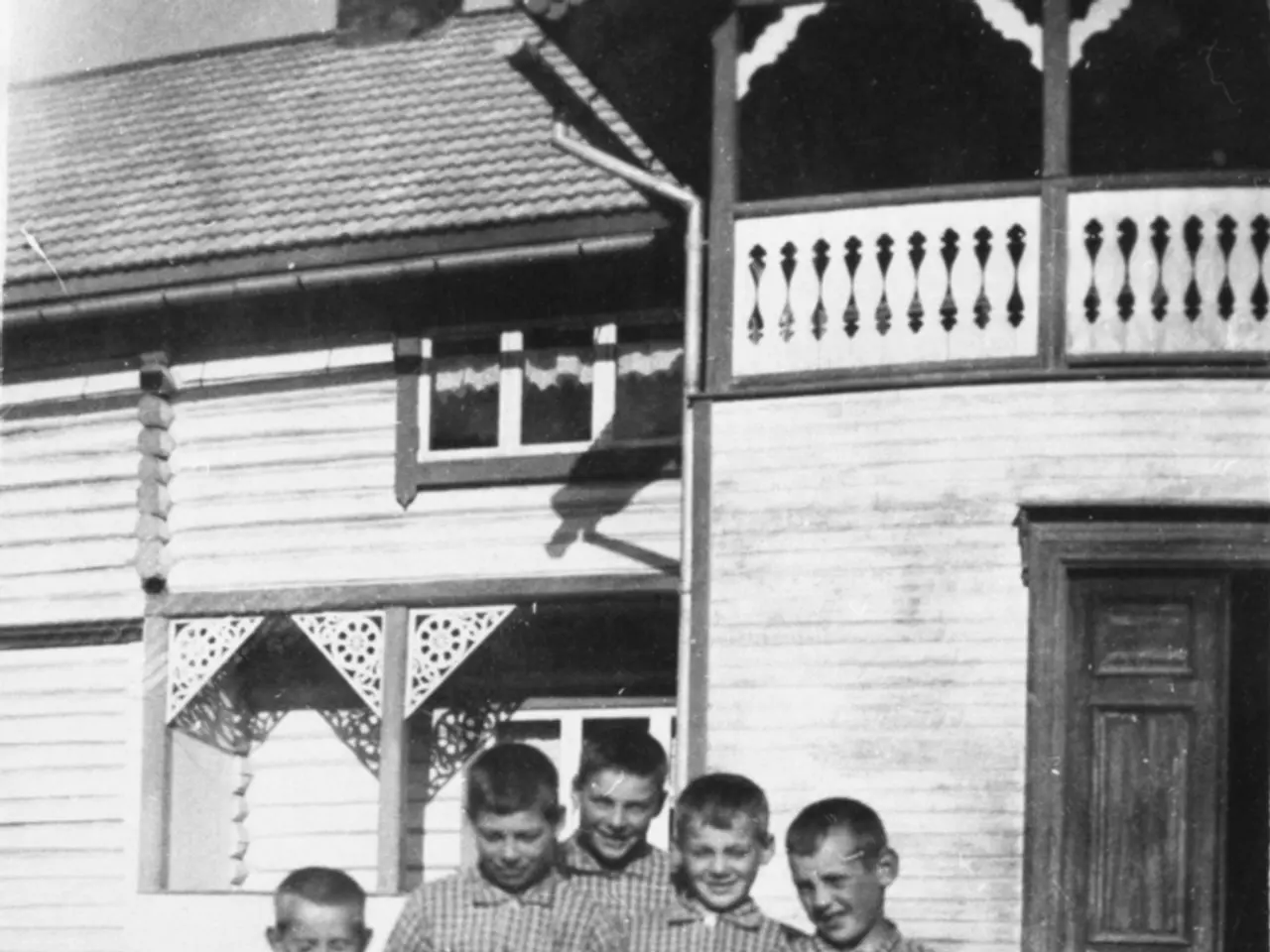 The image shows a black and white photo of a group of children standing in front of a house, with a...