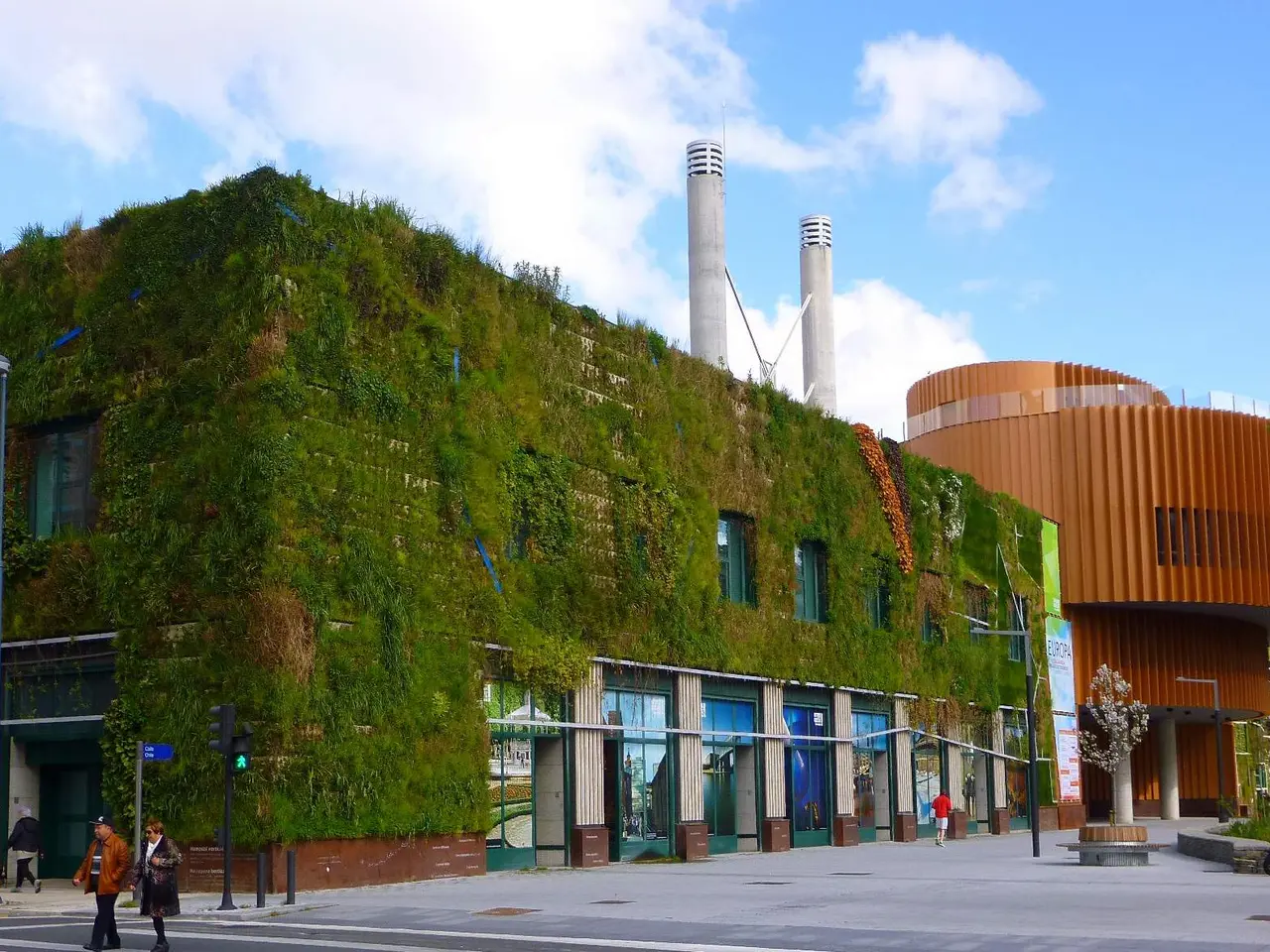 The image shows a large building with a green wall on the side of it, surrounded by street poles,...