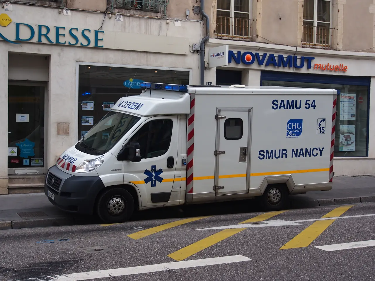 The image shows an ambulance parked on the side of the road in front of a building with windows,...