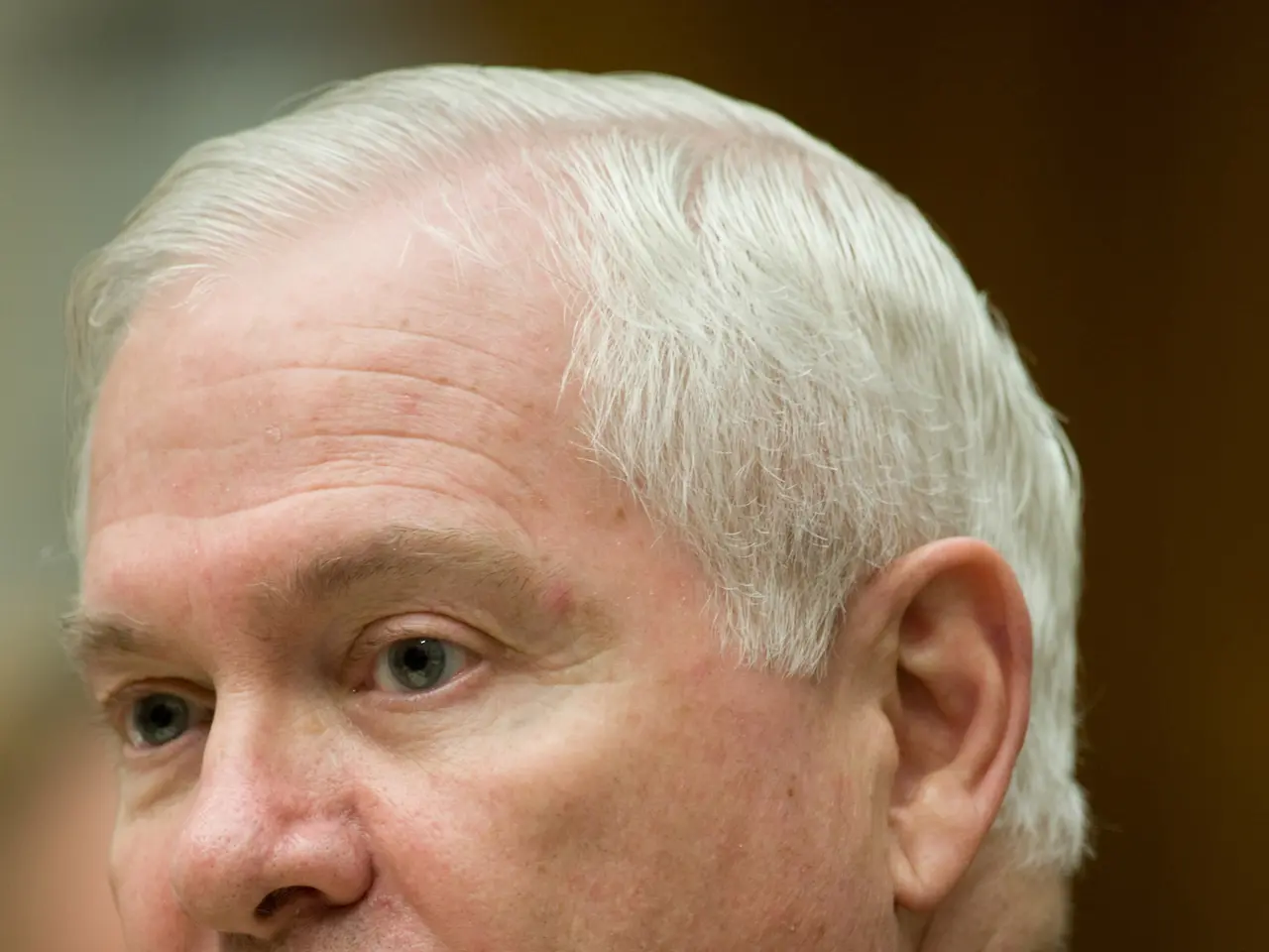 The image shows a man in a black blazer, white shirt, and red tie sitting at a table with a blurred...