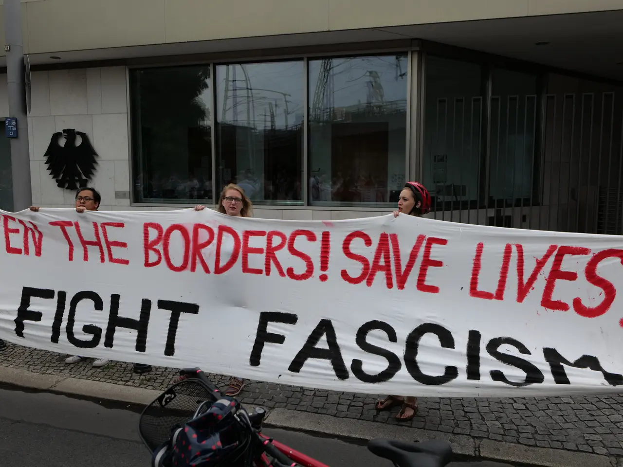 The image shows a group of people holding a banner that reads "Open the Borders, Save Lives, Fight...