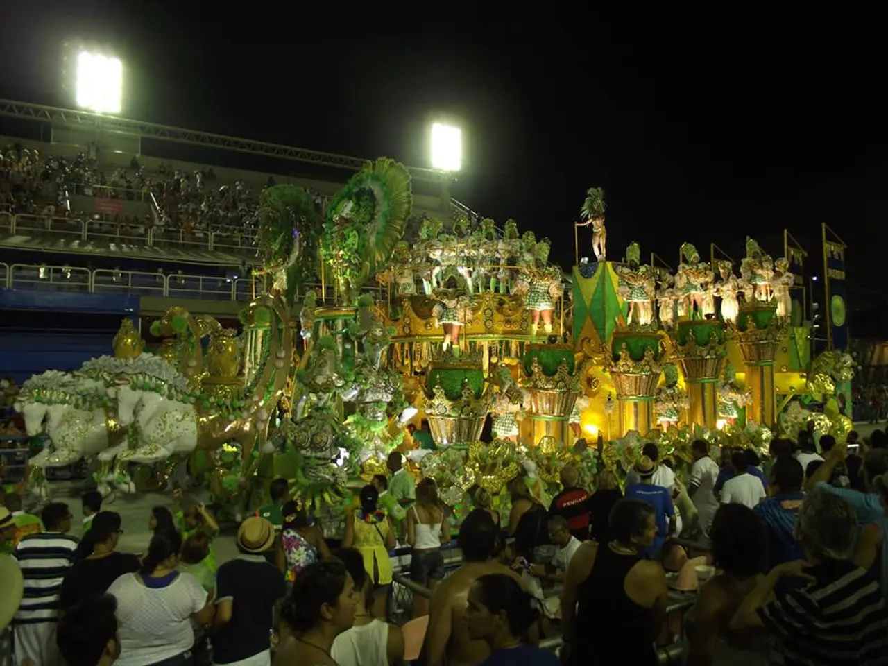 The image shows a large crowd of people standing around a float in a carnival parade at night. The...