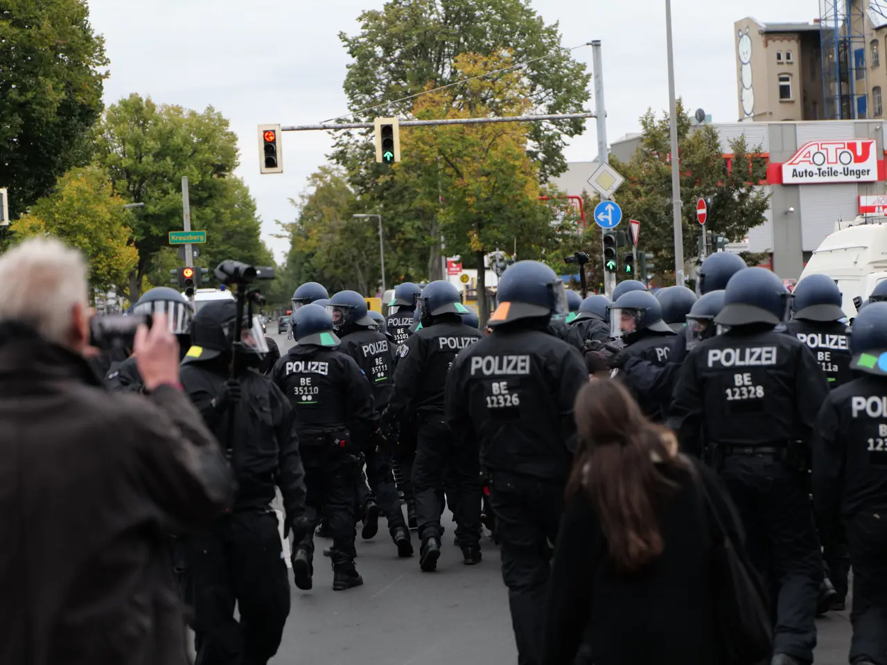 The image shows a group of police officers walking down a street next to a crowd. They are wearing...