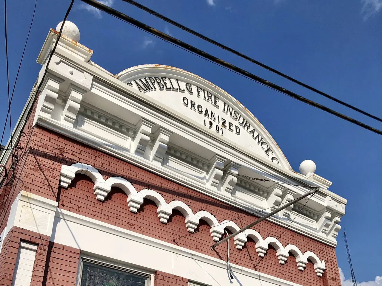 The image shows a red brick building with a white door and windows, a metal fence, some plants, a...