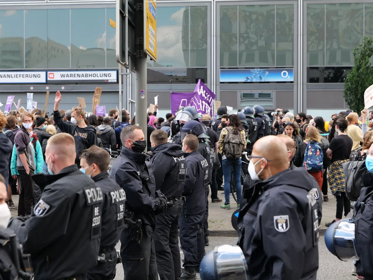 The image shows a large group of people standing in front of a building, some of them holding...