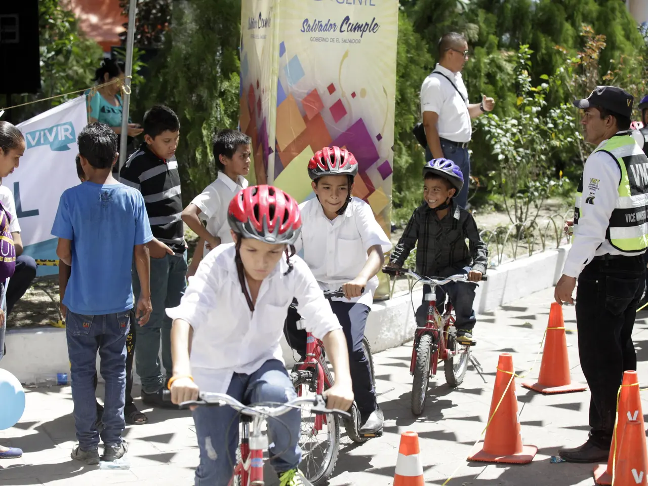 The image shows a group of children riding bicycles down a street lined with traffic cones. Some of...