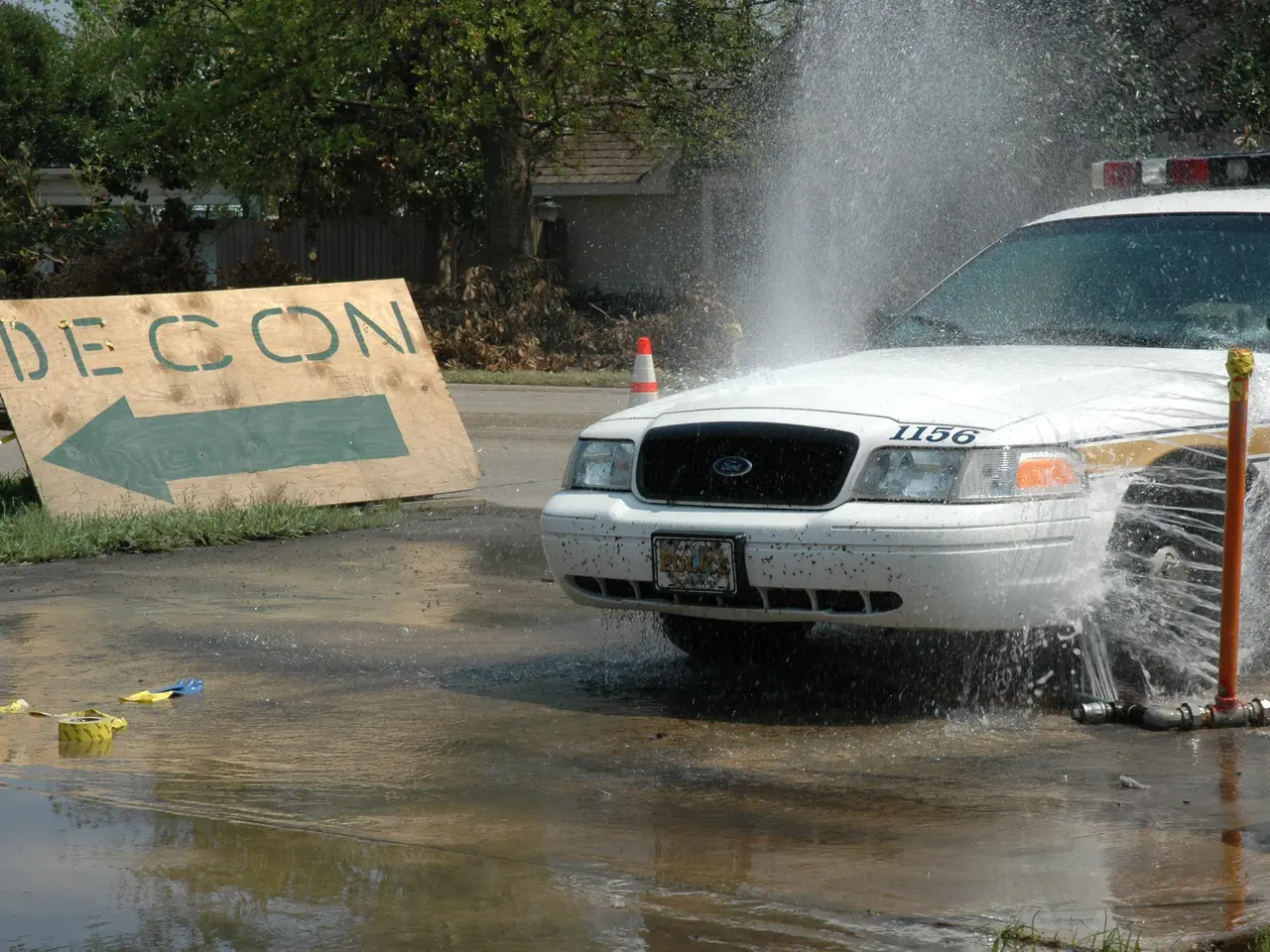 The image shows a police car driving through a puddle of water, with a board with text on it, a...