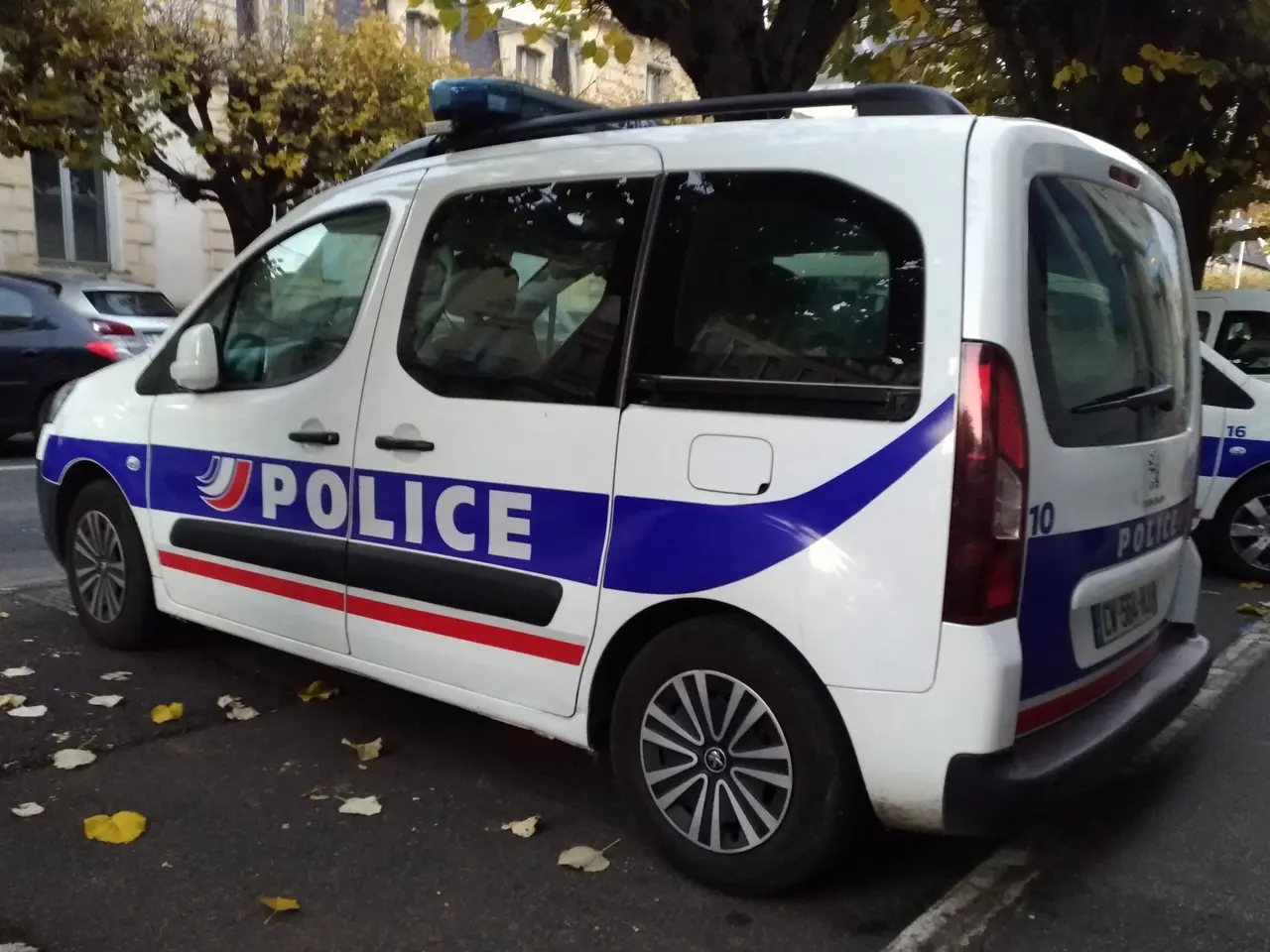 The image shows a police van parked on the side of a street, surrounded by buildings with windows,...