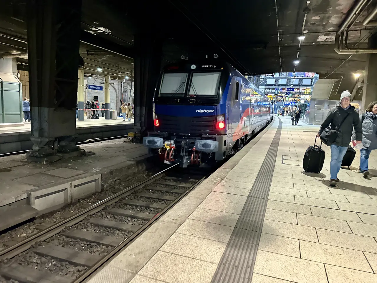 The image shows a train pulling into a train station at night. There are people walking on the...