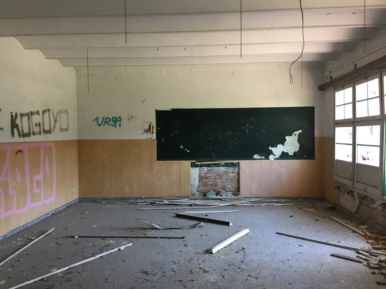 The image shows an empty classroom with graffiti on the walls and a blackboard in the center. On...