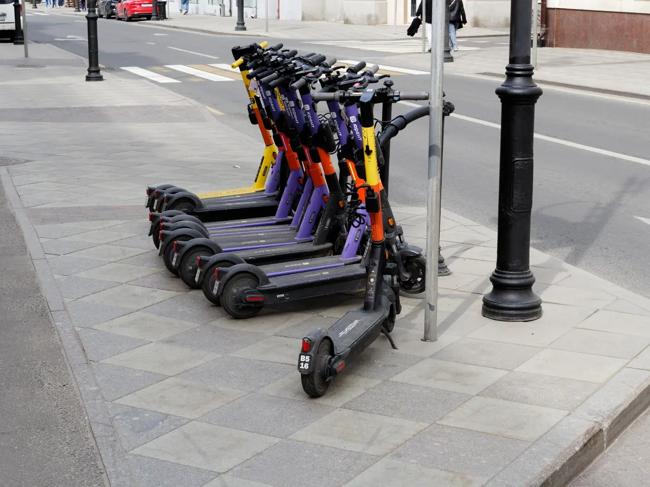 The image shows a row of electric scooters parked on the side of a street, with vehicles driving...