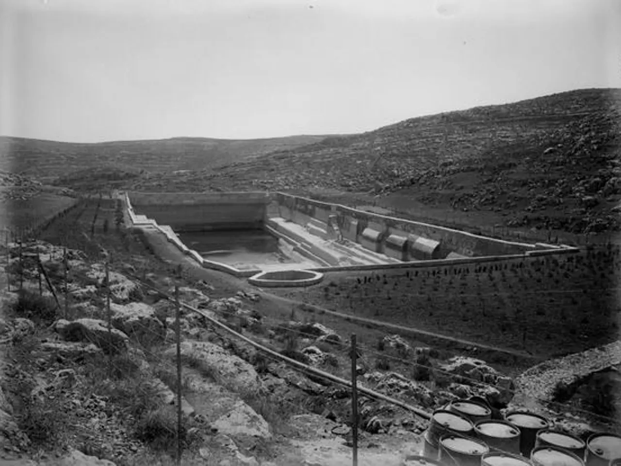 The image shows a black and white photo of a large dam in the middle of a field, surrounded by...