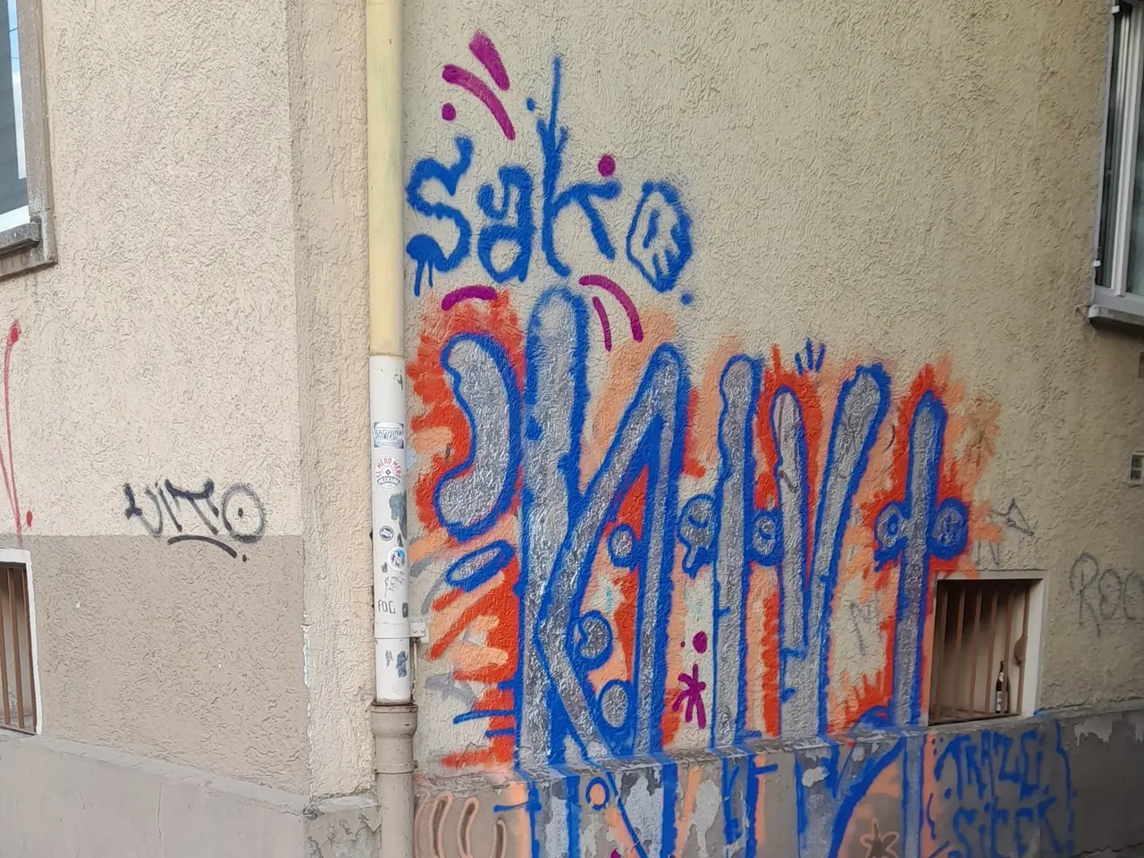 The image shows a building with windows, a pipe, and graffiti on the side of it. At the bottom of...