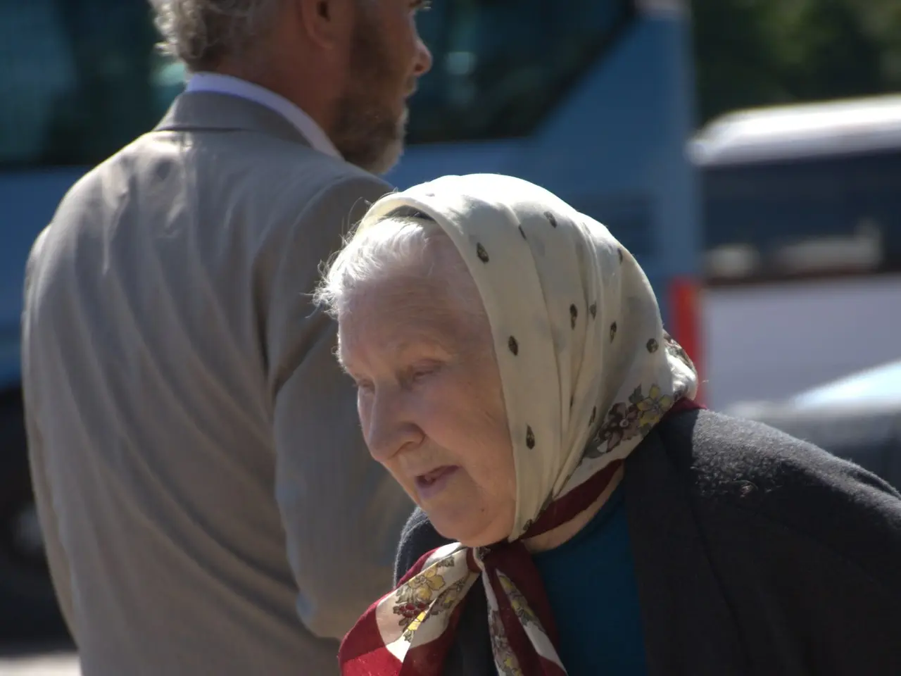 The image shows an elderly woman walking down the street with a cane, accompanied by a man. In the...