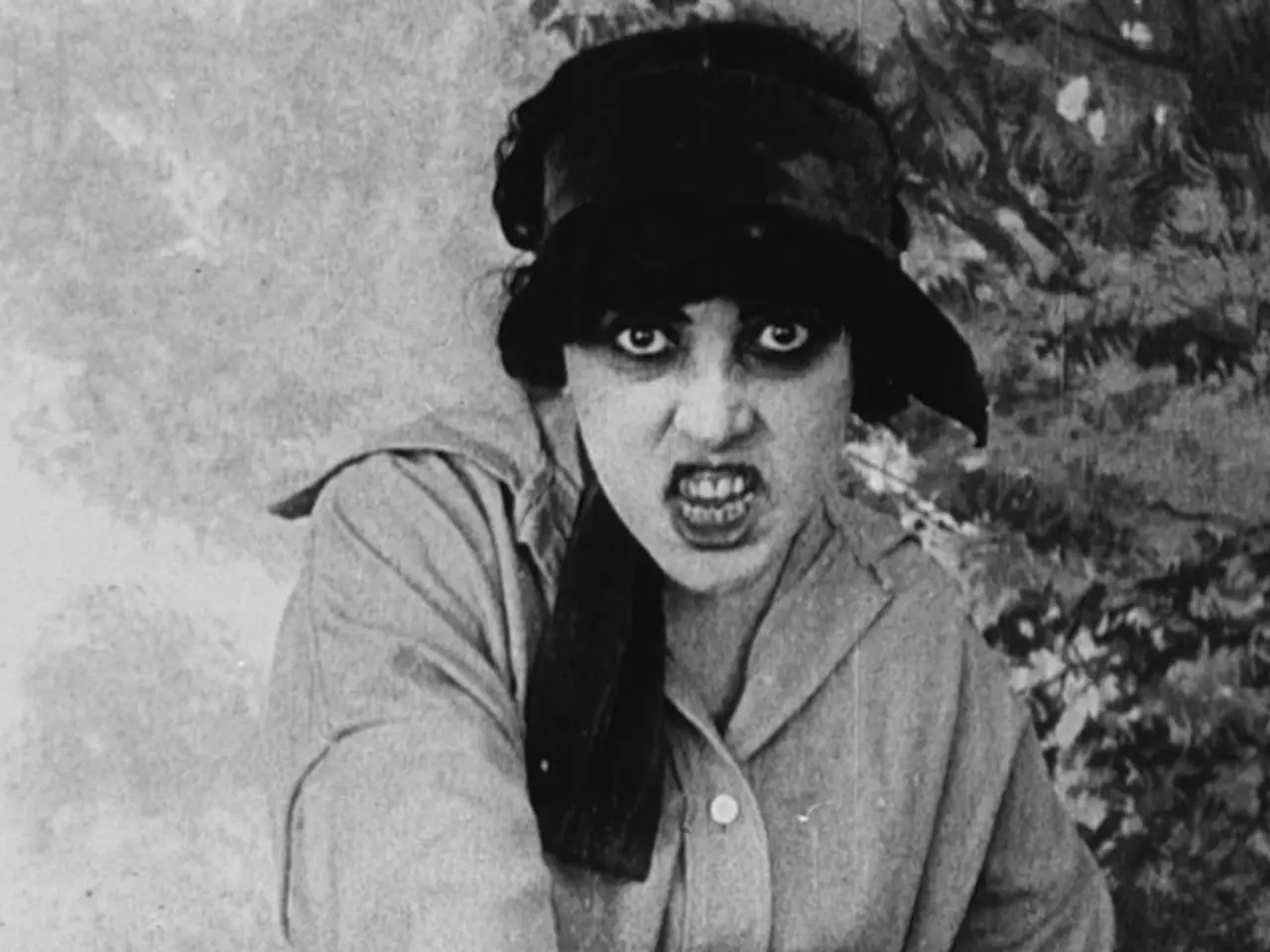 The image shows a black and white photo of a woman wearing a hat, standing in front of a wall with...