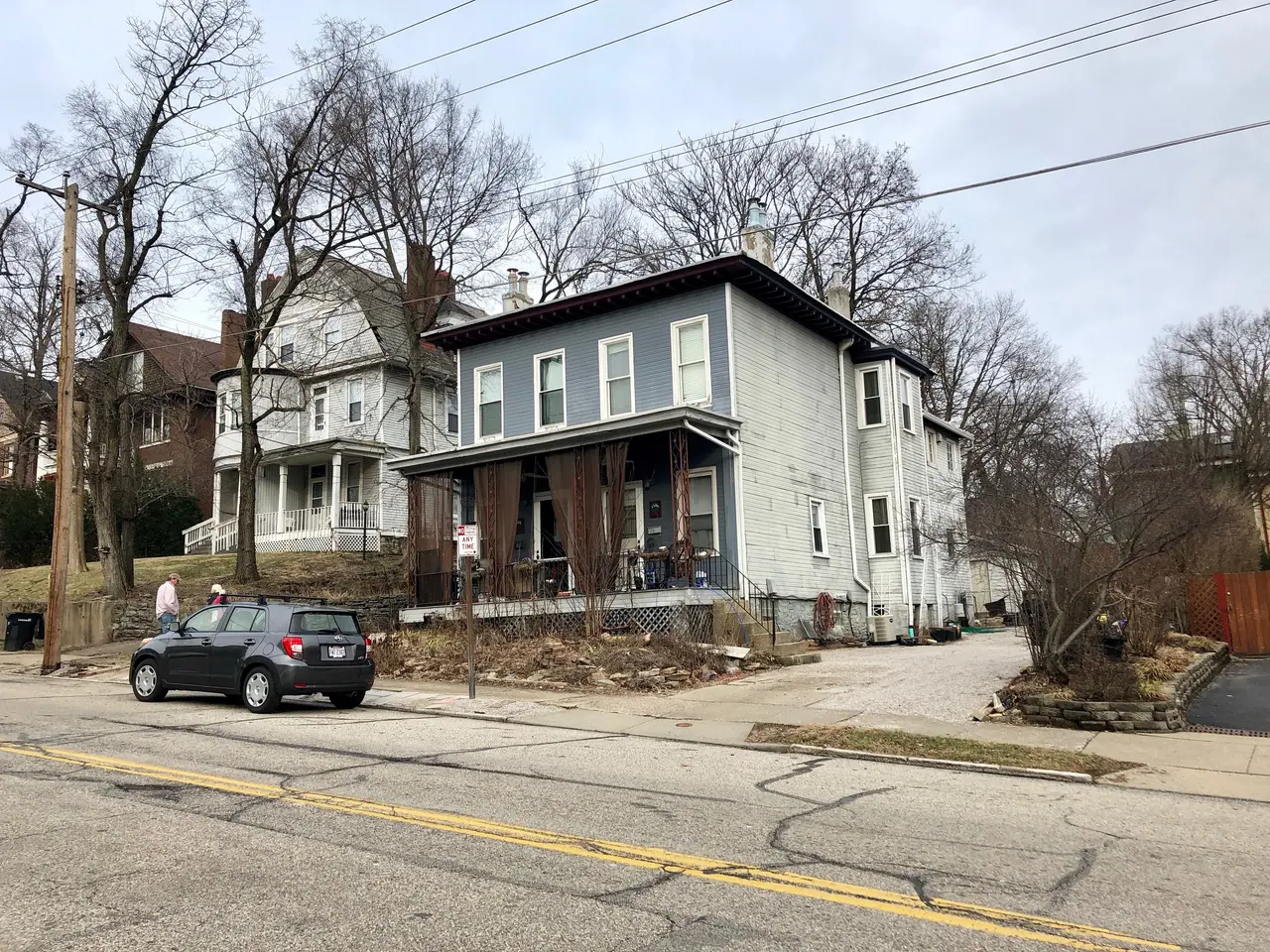 The image shows a car parked on the side of a street next to a row of houses with windows, trees,...