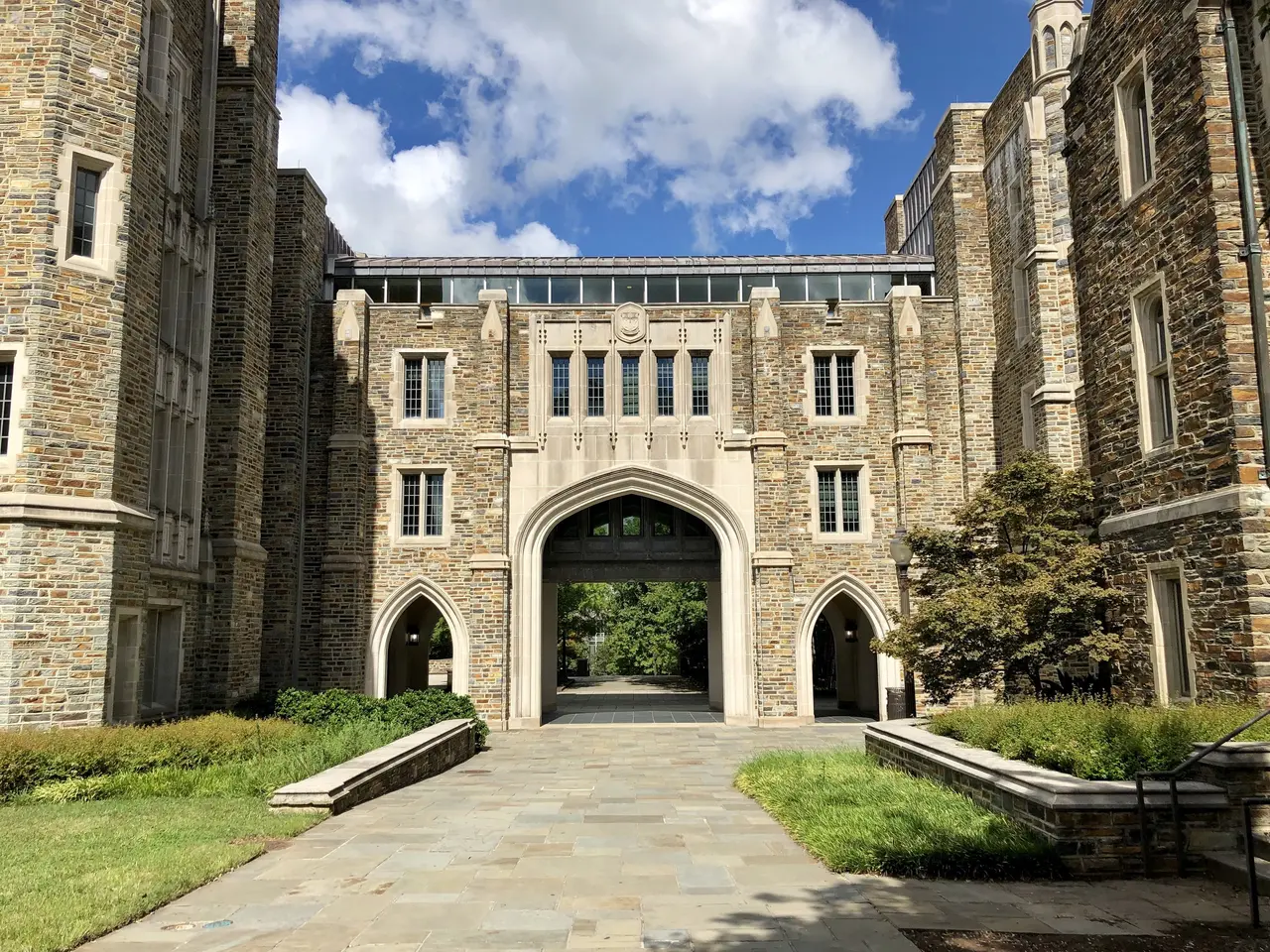 The image shows a large stone building with a walkway in front of it, surrounded by lush green...
