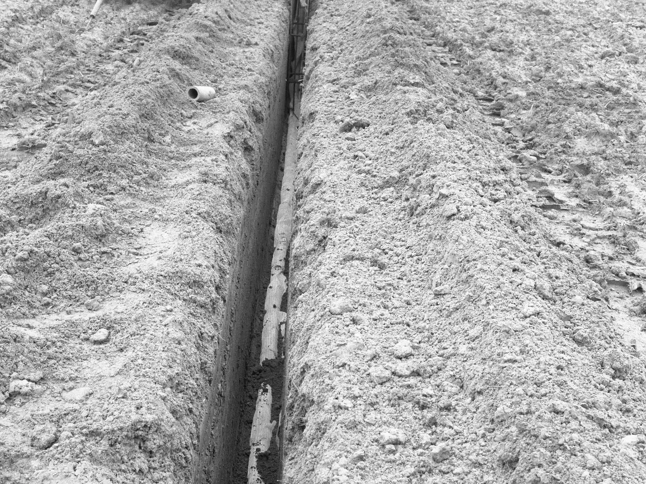 The image shows a black and white photo of a trench dug into the ground, with a pipe running along...