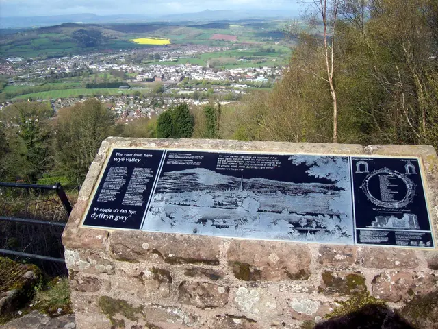 In the image I can see few trees, houses, mountains, fencing and the board is attached to the wall....