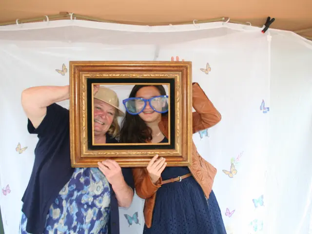In this image we can see two women standing holding a frame. On the backside we can see a curtain...