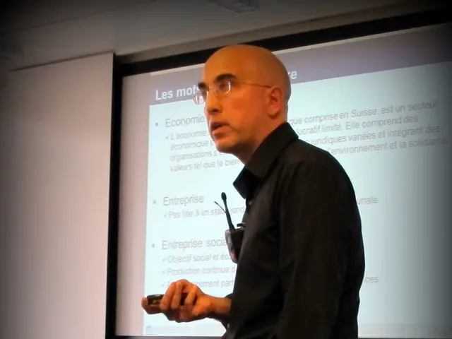 In this picture a guy with a electronic device in his hand and a screen is in the background.