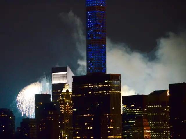 In this image there are buildings and we can see lights. In the background there are fireworks and...