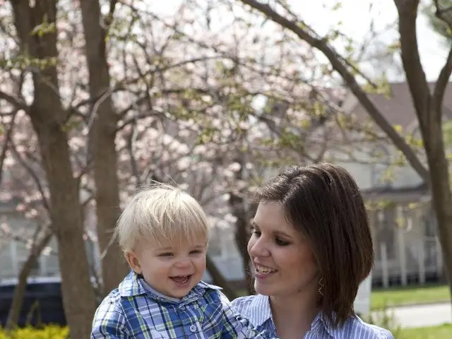 In this image, we can see a woman is carrying a baby. They both are smiling. Background we can see...