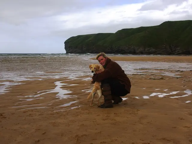 In this picture I can see person holding dog in a sea shore, side we can see hill.