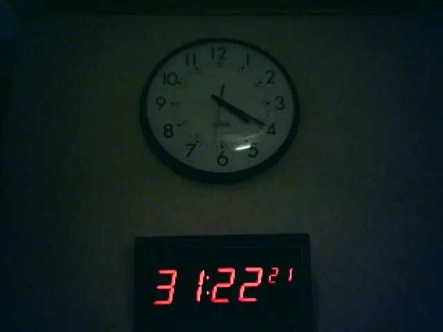 In this image there is a digital clock, above the digital clock there is a wall clock on the wall.