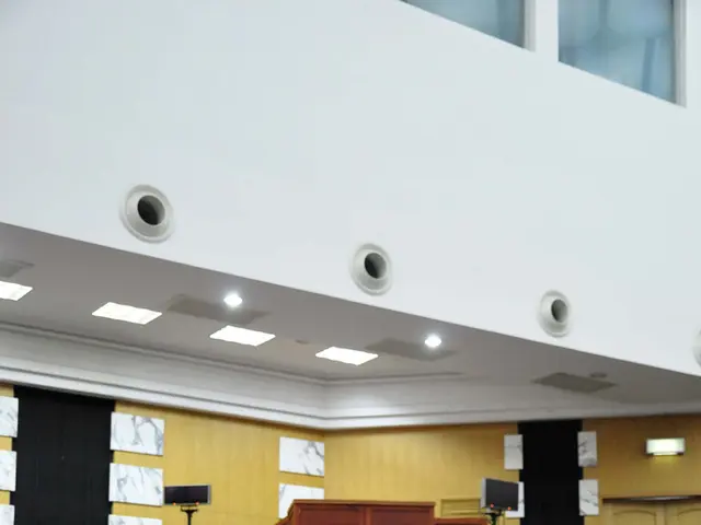 This is looking like a meeting hall. Many people are sitting on chairs. In front of them on table...