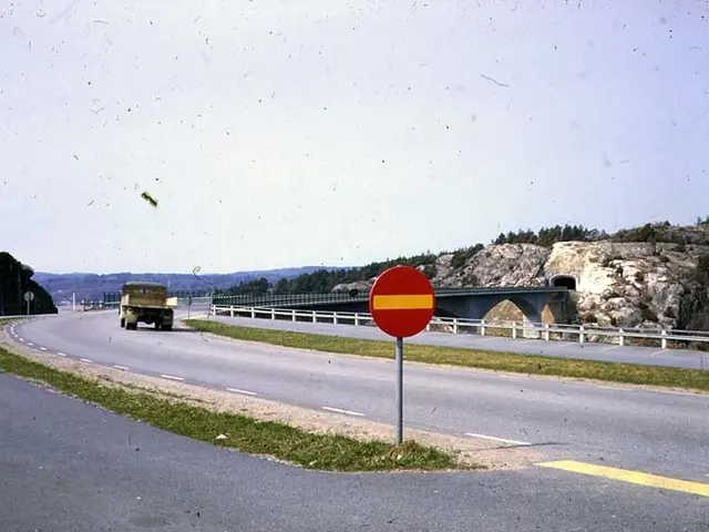 The image shows a truck driving down a road next to a bridge, with a stop sign in the foreground....