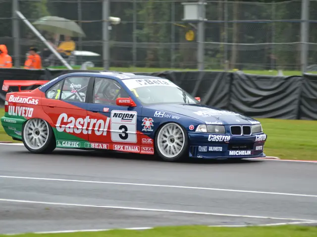 The image shows a car driving on a race track with text and numbers on it, surrounded by grass on...