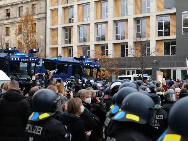 The image shows a large group of people standing in front of a line of police officers wearing...
