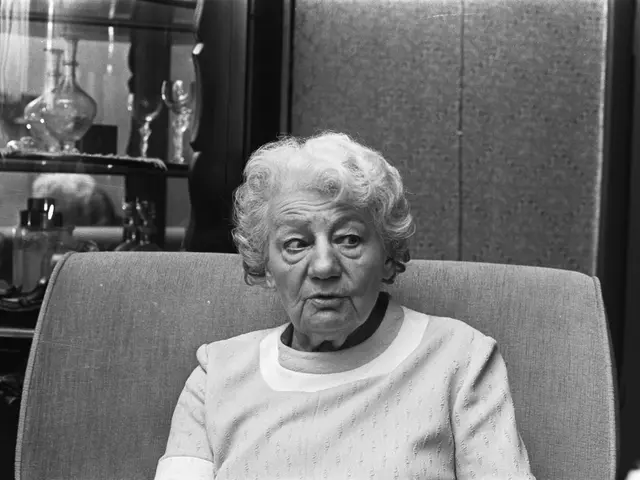 The image shows an elderly woman sitting in a chair in front of a glass display case filled with...