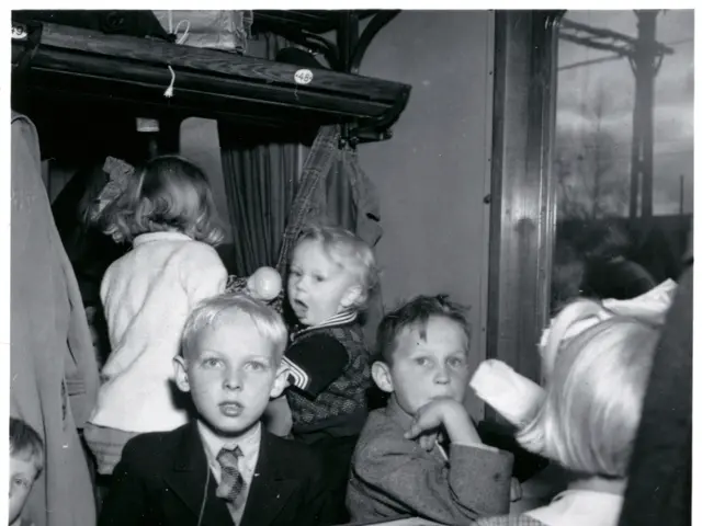 The image shows a black and white photo of a group of children sitting on a train. In the center of...
