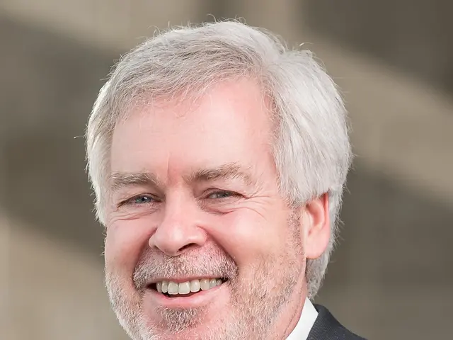 The image shows a man in a suit smiling at the camera with a blurred background. He appears to be...