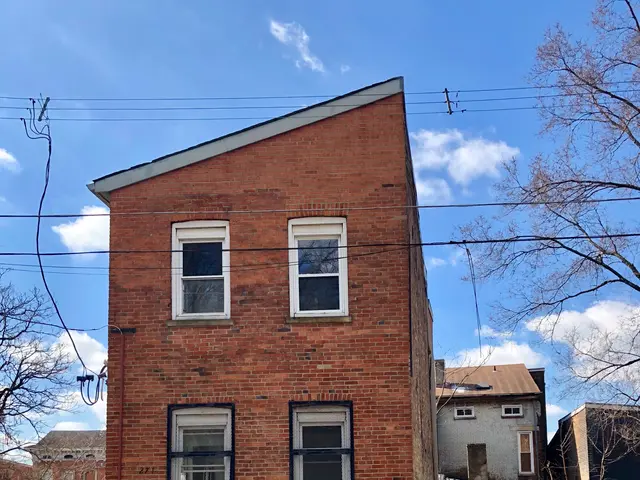 The image shows a red brick building on the corner of a street, with windows, a metal fence, a...