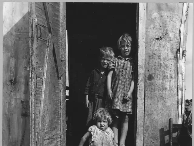 The image shows a black and white photo of three children standing in the doorway of a shack. On...