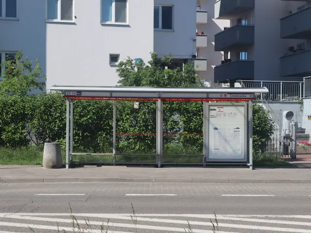 The image shows a bus stop on the side of a road, surrounded by buildings with windows, trees,...
