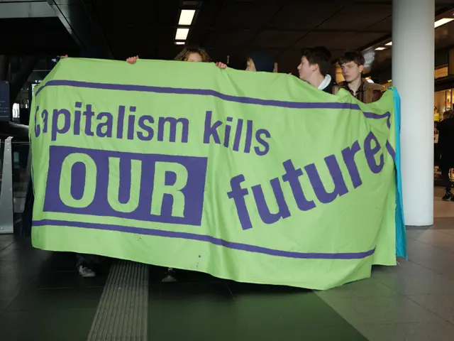 The image shows a group of people standing in an airport, holding a banner that reads "Capitalism...
