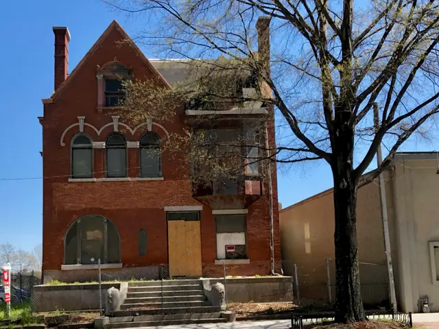 The image shows a red brick house with a tree in front of it, surrounded by a metal fence, with a...