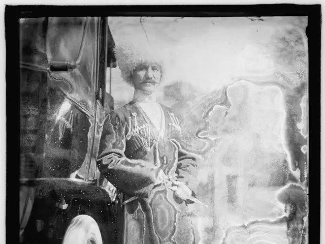 The image shows an old black and white photo of a man standing next to a car. He is wearing a long...