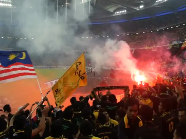 The image shows a large crowd of people in a stadium with flares in the air, some of them holding...