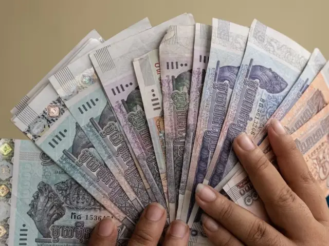 The image shows a person holding a bunch of Thai money in their hands, with a blurred background...