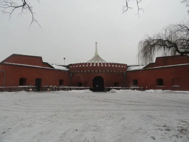 The image shows a large brick building with a dome on top of it, surrounded by a fence, with snow...